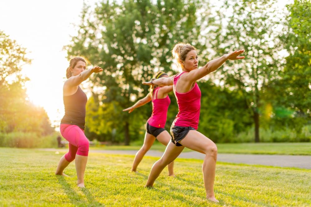 horizontal group of women doing the warrior yoga pose outdoors at sunset with lens flare